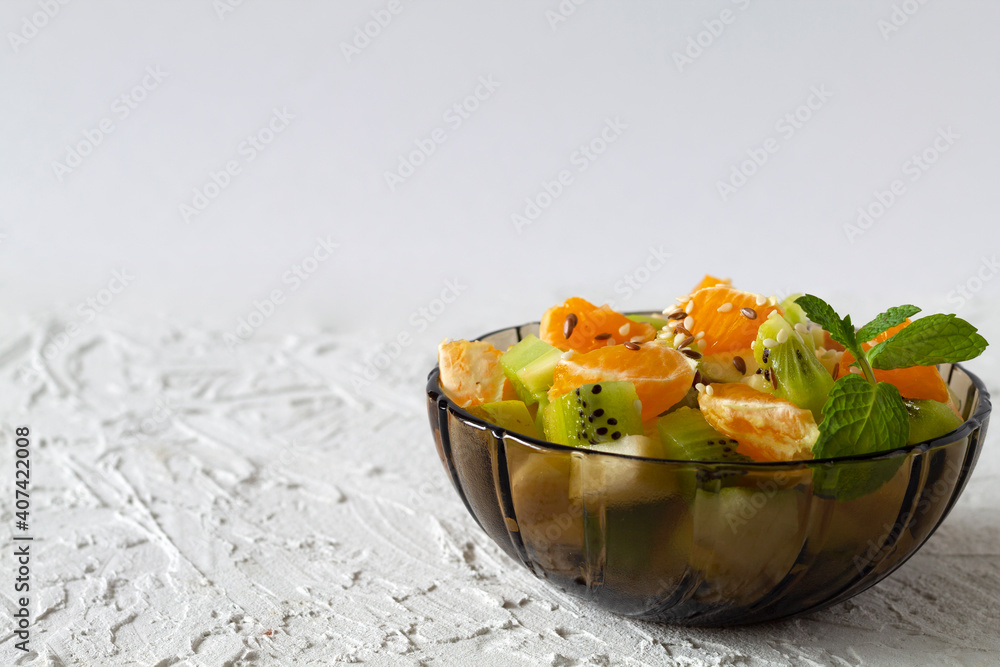 Healthy vegan lunch. Bowl of healthy salad from fruit with green mint on a white table. Mixed fresh banana, orange, kiwi, fruit. copy space. fruit salad in glass dark bowl on white background