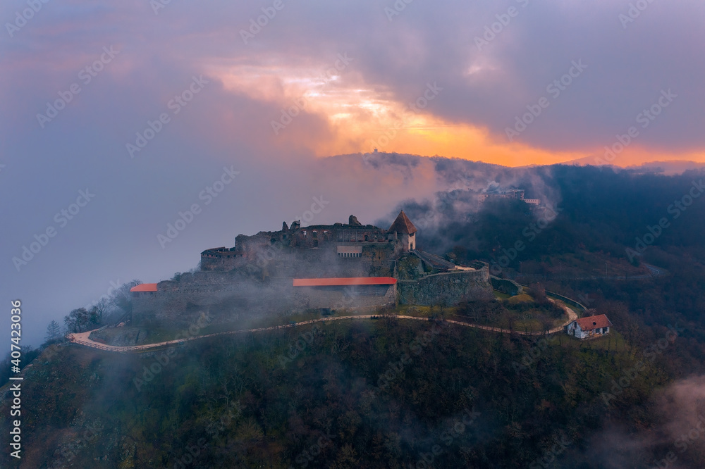 Visegrad, Hungary - Aerial panoramic drone view of the beautiful high castle of Visegrad on a moody winter morning. Danube Bend (Dunakanyar) under fog and amazing golden sunrise at background. Visegrad, Hungary - Aerial panoramic drone view of the beautiful high castle of Visegrad on a moody winter morning. Danube Bend (Dunakanyar) under fog and amazing golden sunrise at background.
