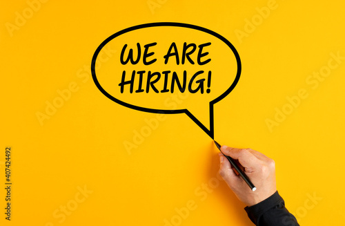 Male hand drawing a speech balloon with the message of we are hiring on yellow background. Job employment or job advert