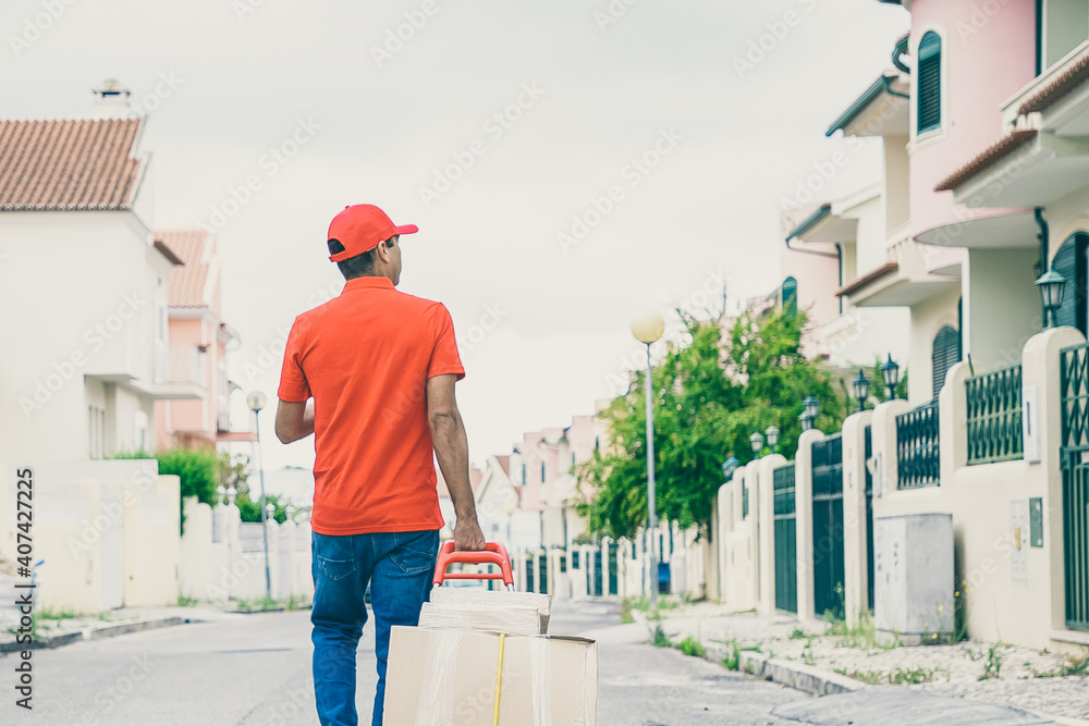 Back view of courier wheeling trolley with carton boxes. Professional courier in red cap and shirt delivering parcels to clients on foot and pacing on street. Delivery service and post concept Back view of courier wheeling trolley with carton boxes. Professional courier in red cap and shirt delivering parcels to clients on foot and pacing on street. Delivery service and post concept