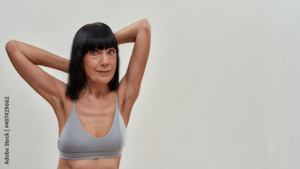 Beautiful half naked caucasian senior woman in lingerie keeping arms raised and looking at camera while standing against grey background