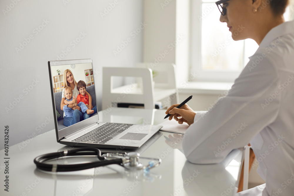 Profile of young female doctor making notes during online consultation with woman and two children patients on laptop from medical clinic office. Telehealth, telemedicine cocnept Profile of young female doctor making notes during online consultation with woman and two children patients on laptop from medical clinic office. Telehealth, telemedicine cocnept