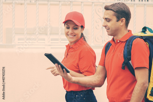 Side view of couriers delivering order and holding tablet. Man and woman wearing jeans and red shirts. Deliveryman carrying thermal backpack. Food delivery service and online shopping concept