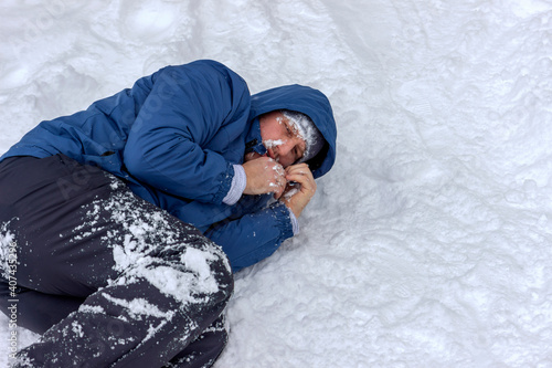 Frozen man in a blue jacket and hat lying down covered snow and frost, trying to stay warm on a very cold winter day, snow falls around him. Sick mountaineer with hypothermia on snow during the day.