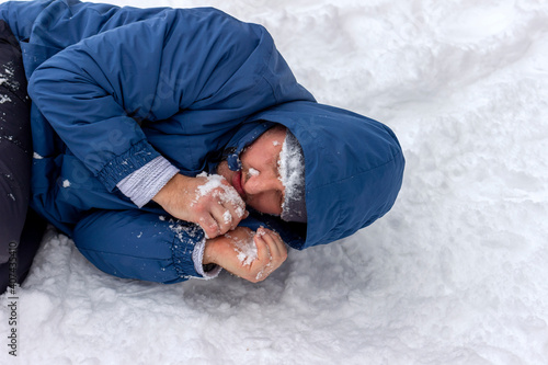 Obraz na plátne Frozen man in a blue jacket and hat lying down covered snow and frost, trying to stay warm on a very cold winter day, snow falls around him