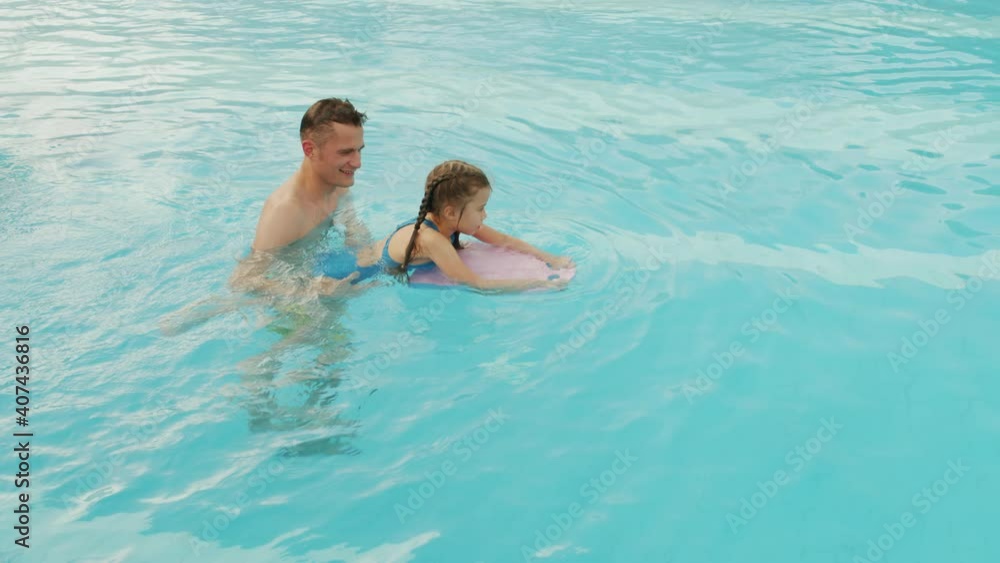 Slow-motion from-above shot of cheerful dad giving swimming lessons to his little daughter with kickboard in big outdoor thermal pool with crystal clear water Slow-motion from-above shot of cheerful dad giving swimming lessons to his little daughter with kickboard in big outdoor thermal pool with crystal clear water