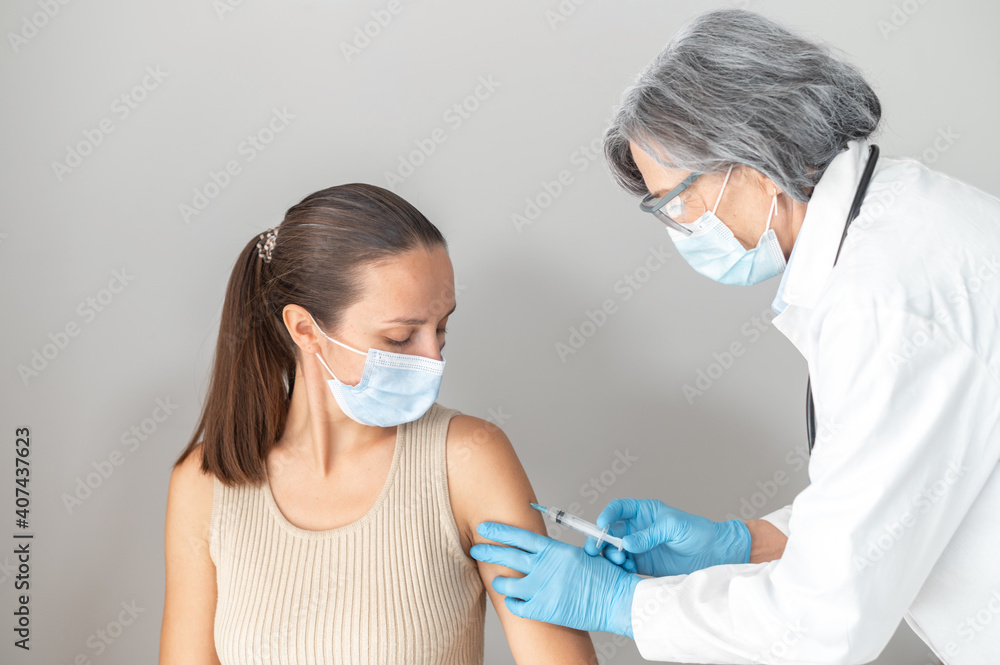 Portrait of two people isolated over gray background. The young europian ethnicity lady volunteered to get a shot of coronavirus vaccine, a senior female doctor in mask injecting flu covid-19 vaccine