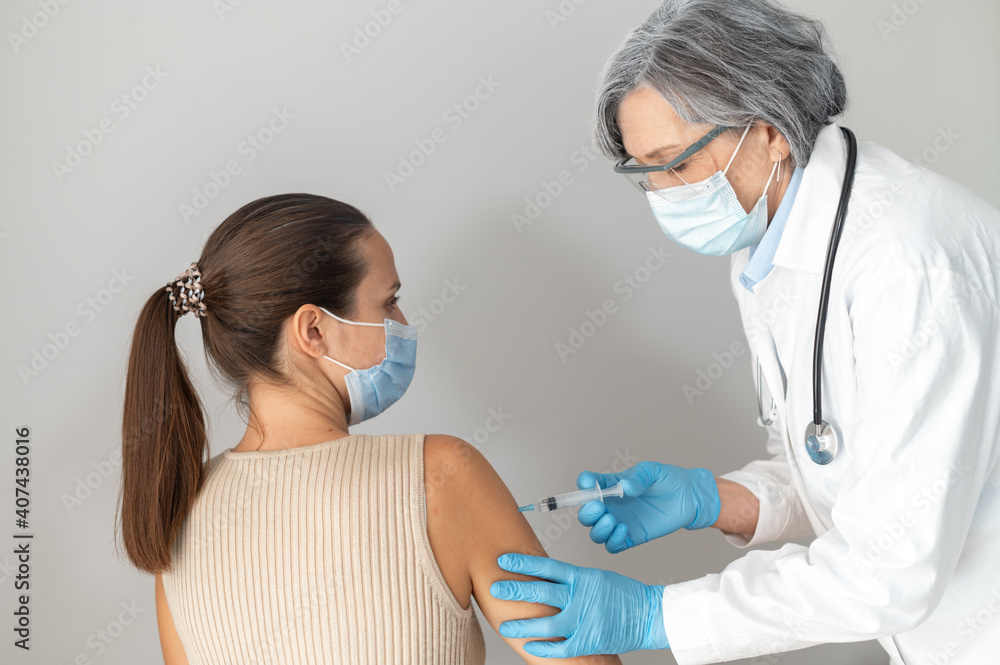 Two people isolated over gray background, senior mature doctor in protective face mask and gloves injecting shot of flu vaccine in the upper-arm. Young lady volunteered for coronavirus immunization Two people isolated over gray background, senior mature doctor in protective face mask and gloves injecting shot of flu vaccine in the upper-arm. Young lady volunteered for coronavirus immunization