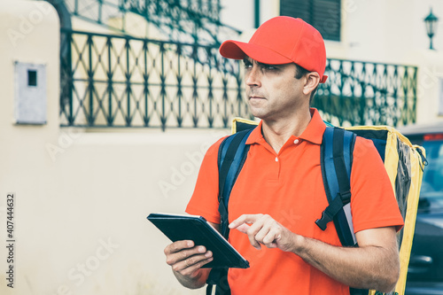 Frowning courier standing and watching address via tablet in hands. Pensive deliveryman delivering order in thermo backpack and wearing red shirt and cap. Delivery service and online shopping concept