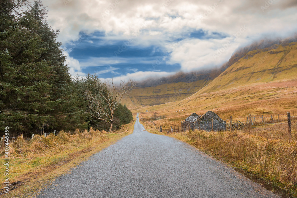 Small road into mountains, Forest on the left, empty fields and small abandoned house on the right. Geniff Horseshoe loop drive, county Sligo, Ireland. Cloudy sky. Nature landscape scene