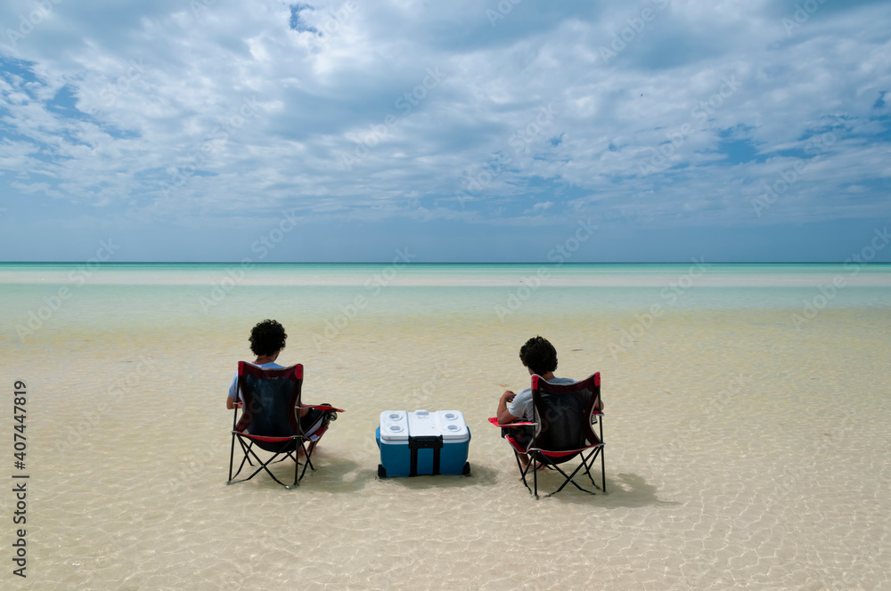 Two guys sitting in the middle of the Caribbean Ocean on chairs and iceboxes, watching the horizon. Holbox Island, Mexico. An ideal place to relax