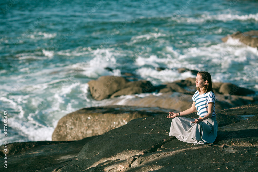 Young yoga woman meditates in the lotus position sitting on rocks on the Atlantic ocean coast.