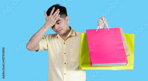 Portrait handsome young asian man wearing yellow stressful content guy carrying new things colorful packages on blue background in studio. Asian man people. photo