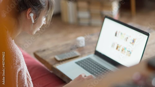 young unrecognizable woman with wireless earphones sits at wooden desk at home, browsing online store on laptop and making online order from home by phone. focus on earphone