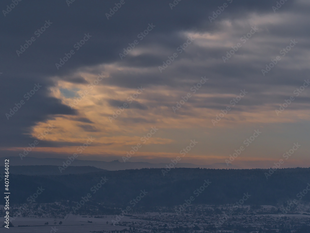 Beautiful aerial panoramic view of foothills of Swabian Alb, Germany in winter with village, snow-covered fields and silhouettes of Hohenzollern Castle below dramatic clearing sky of orange clouds.