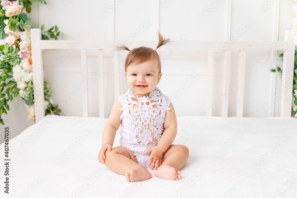 cheerful baby girl six months old sitting in a bright beautiful room on a white bed in a lace bodysuit and smiling cheerful baby girl six months old sitting in a bright beautiful room on a white bed in a lace bodysuit and smiling