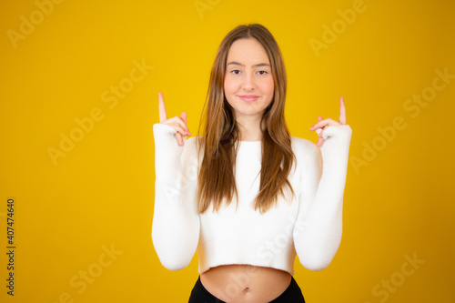 Happy beautiful young woman in white sweater is pointing up, looking away and talking. Waist up studio shot on yellow background.