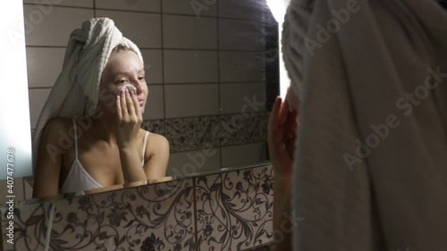 woman with a towel on her head in the bathroom after a shower looks in the mirror and cleans the skin with a cotton swab. 