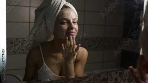 Beautiful caucasian woman with a towel on her head in the bathroom applies cream after a shower to her face and smiles. 