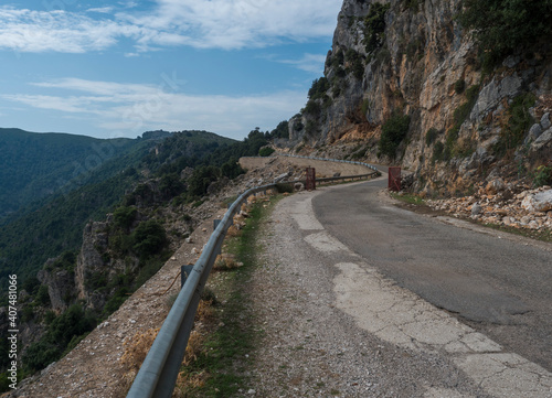 Narrow dangerous asphalt road at Genna Croce pass. at Supramonte Mountains with white limestone rocks, green hills, trees and mediterranean forest vegetation. Sardinia, Italy. Summer cloudy sky.