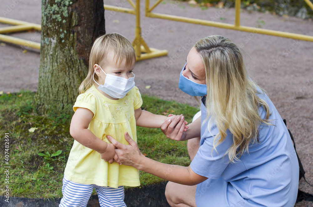 Fototapeta premium Woman and child girl in the park wearing medical face mask. Mother and her little daughter in surgical mask, closed park for visitors. Wearing surgical masks in public.