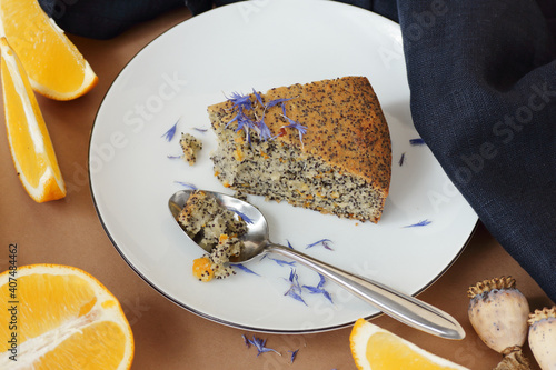 Poppy seed and orange peel vegan egg free and dairy free cake, bite on the orange plate with blue linen textile and poppy head nearby, closeup, copy space, vegan food and pastry concept photo