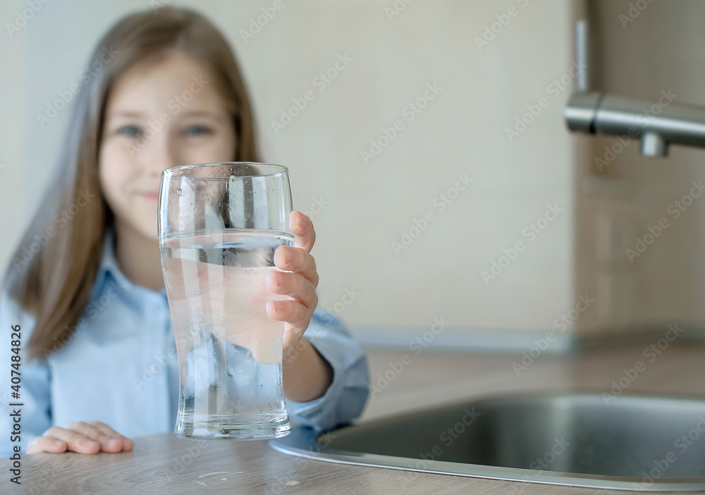 Happy little girl holding a glass of water. Selective focus on child hand with glass of water in foreground. Hydration. Water quality check. World water monitoring day. Environmental pollution problem Happy little girl holding a glass of water. Selective focus on child hand with glass of water in foreground. Hydration. Water quality check. World water monitoring day. Environmental pollution problem