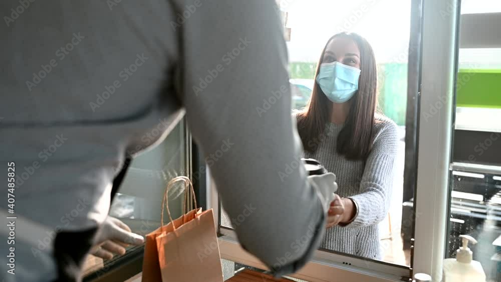 A female customer takes a package with order and a paper cup of hot coffee from a waiter wearing mask and gloves through an improvised take-away window for safety. Cafe works in take out due pandemic A female customer takes a package with order and a paper cup of hot coffee from a waiter wearing mask and gloves through an improvised take-away window for safety. Cafe works in take out due pandemic