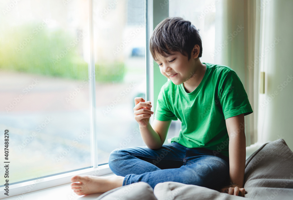 Authentic portrait of Happy Kid with smiling face siting next to window with morning light, Active boy looking at his fingers with thinking face, Child self isolation at home during covid-19 lockdown. Authentic portrait of Happy Kid with smiling face siting next to window with morning light, Active boy looking at his fingers with thinking face, Child self isolation at home during covid-19 lockdown.