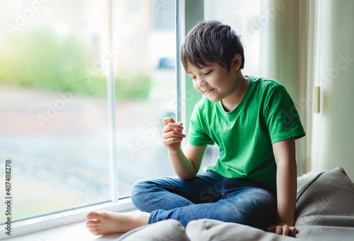 Free Download Authentic portrait of Happy Kid with smiling face siting next to window with morning light, Active boy looking at his fingers with thinking face, Child self isolation at home during covid-19 lockdown. - GFXLoad