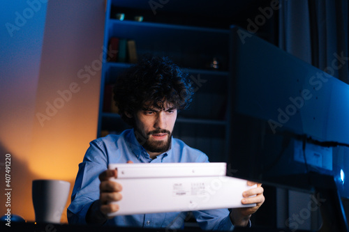 Bearded young freelancer shaking wireless keyboard while working on computer sitting on desk at home office workplace at late night in dark room. Concept of remote working, distance learning, photo