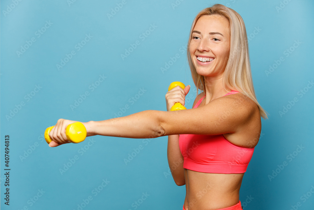 Portrait of a smiling fit young sportswoman. Positive fitness woman holding dumbells over blue background. Portrait of a smiling fit young sportswoman. Positive fitness woman holding dumbells over blue background.