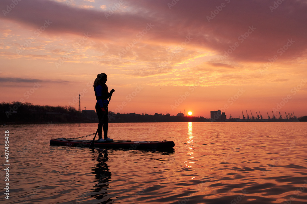 Silhouette of a young woman standing on a SUP during a beautiful winter sunrise on the river