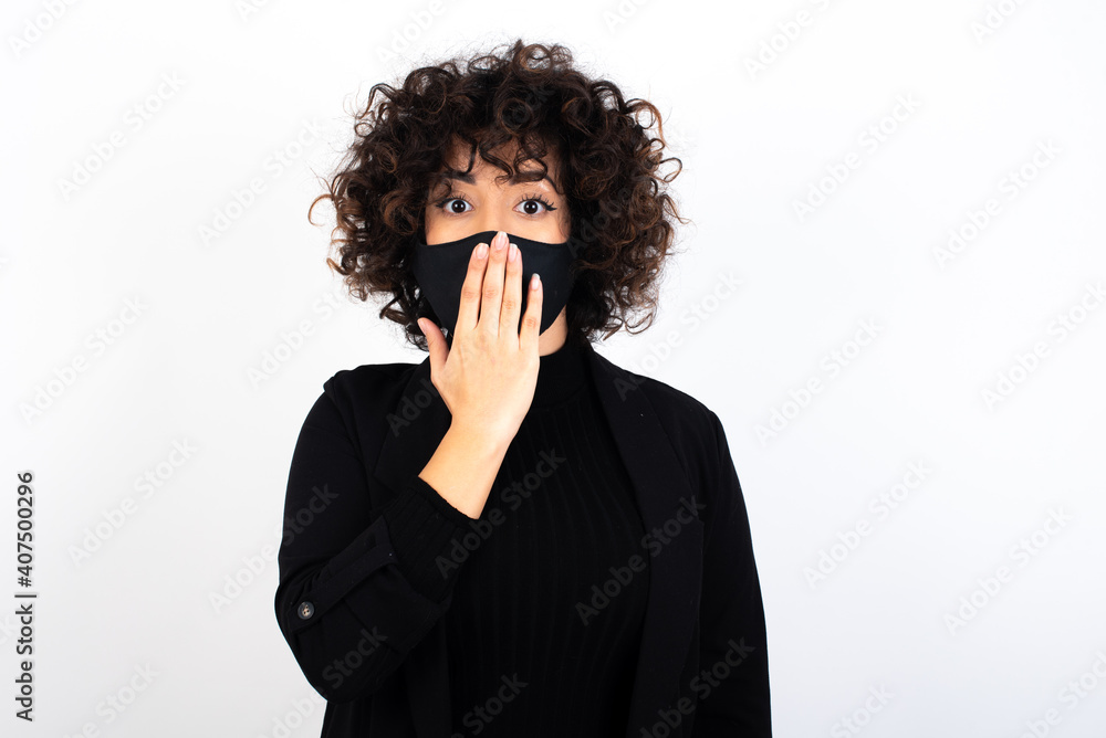 Oh! I think I said it! Close up portrait young beautiful caucasian woman wearing medical mask standing against white wall, cover open mouth by hand palm, look at camera with big eyes. Oh! I think I said it! Close up portrait young beautiful caucasian woman wearing medical mask standing against white wall, cover open mouth by hand palm, look at camera with big eyes.