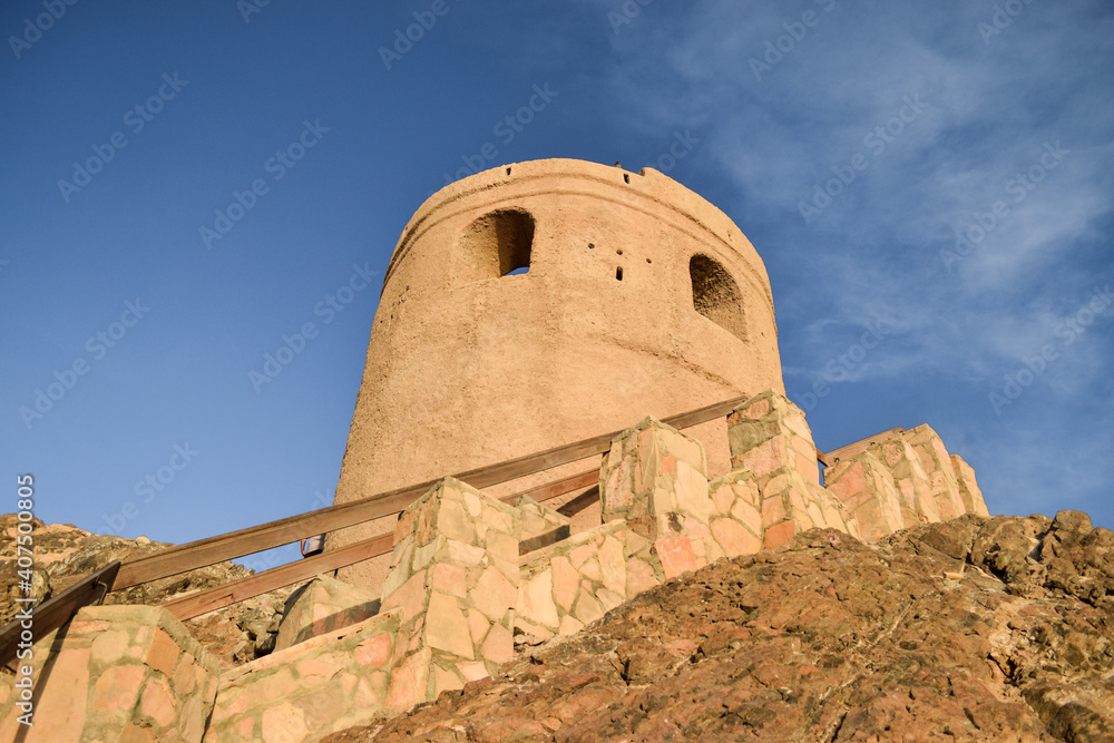 Bottom-up view of an ancient watch tower on the top of a hill in Muscat, Oman, in contrast with the blue sky.