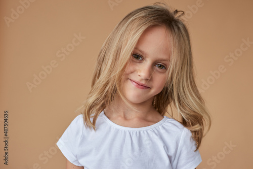 Closeup front portrait of pretty little girl with wavy blond hair, dressed in white clothes, over beige background.
