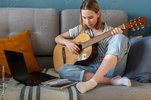 Online music learning. Girl playing acoustic guitar and watching online course on laptop while lesson at home. Online training, online classes. Remote training.Online education