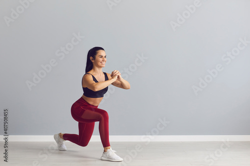 Smiling fitness trainer showing how to do fitness exercise. Happy fit young woman in sports bra and leggings doing forward lunges holding hands together in front of chest during workout at the gym photo