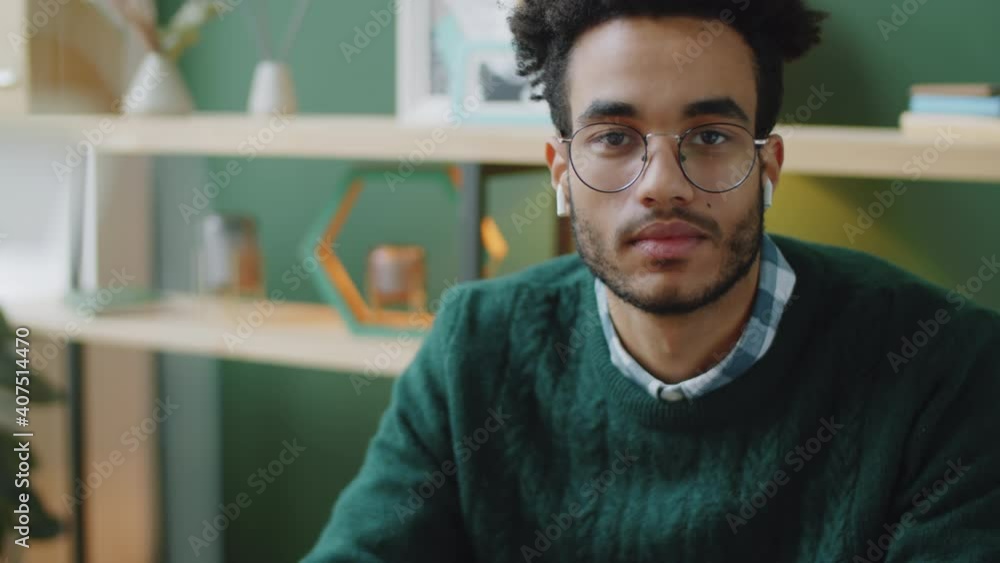Portrait of young handsome mixed race businessman in wireless earphones sitting at workplace in office and looking at camera Portrait of young handsome mixed race businessman in wireless earphones sitting at workplace in office and looking at camera