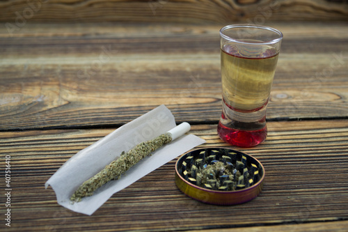 Joint, grinder, cannabis buds, alcohol shot and related items on a table.
