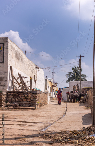 Lakundi, Karnataka, India - November 6, 2013: Typical side street with central gutter and white painted houses under blue cloudscape. Trash and black cows around. Walking woman in sari. photo