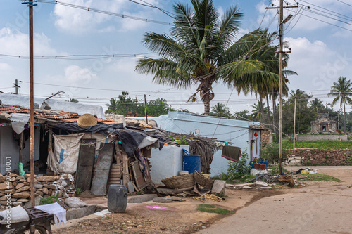 Lakundi, Karnataka, India - November 6, 2013: Couple of poor houses on street corner with lots of trash and stuff around under blue cloudscape and tall green palm tree. photo