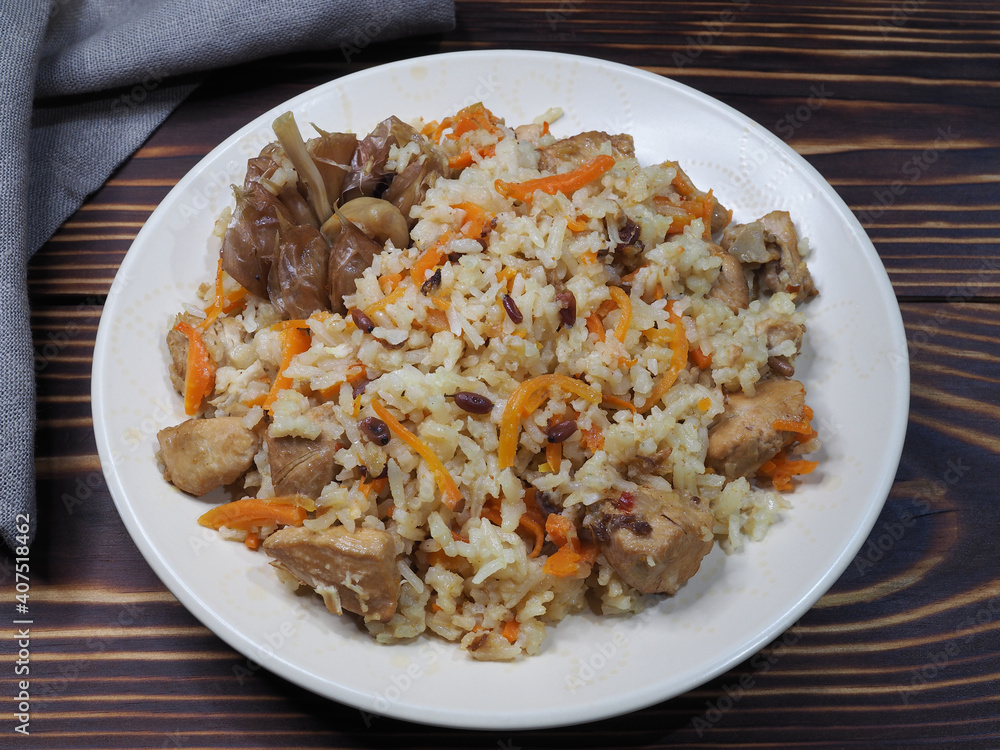 Traditional national dish pilaf. Garlic, carrots, rice, meat with seasonings and sauces in a plate on a wooden table, flat layout. Delicious Uzbek hot food plov with chicken, cereals and vegetables Traditional national dish pilaf. Garlic, carrots, rice, meat with seasonings and sauces in a plate on a wooden table, flat layout. Delicious Uzbek hot food plov with chicken, cereals and vegetables