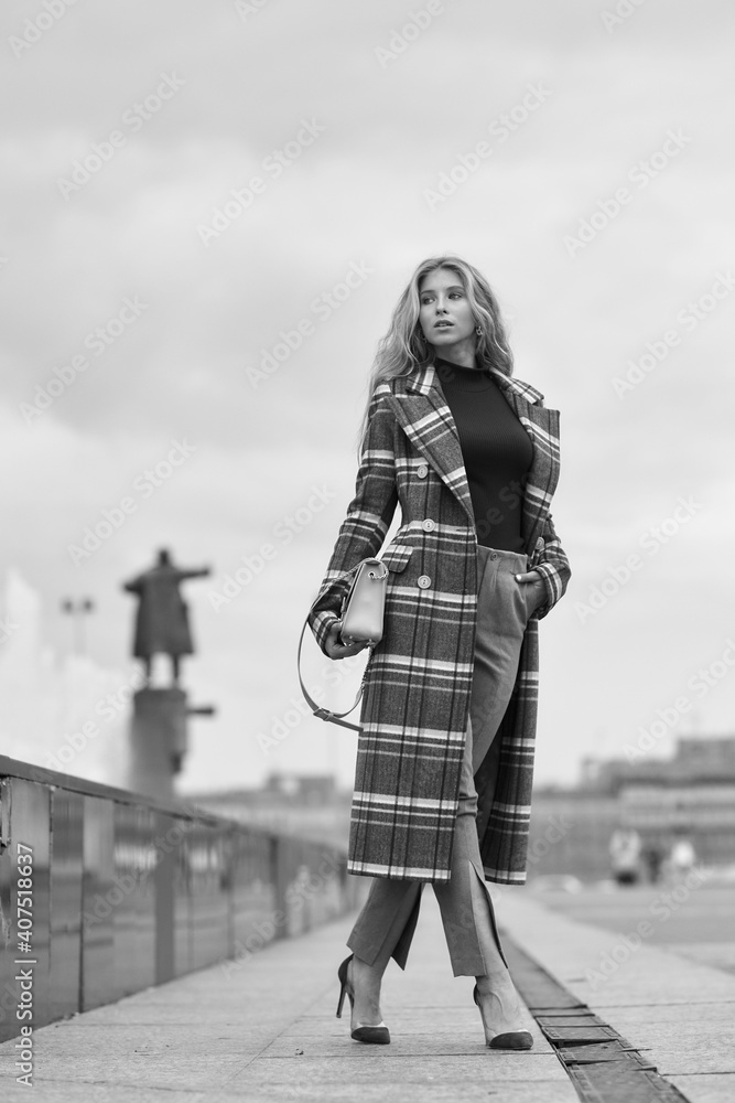 Elegant woman walking city street. Fashion fall autumn look. Pretty caucasian blonde stylish girl wearing blue jeans, wine red pullover and squared wool coat. Female with makeup and wavy blonde hair