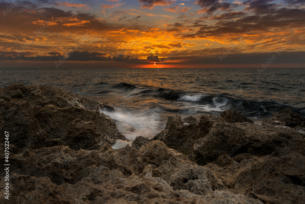 Fototapeta premium Red sunset, just swallowed by the sea. Warm colors like orange, red and yellow. The waves of the Mediterranean sea that crash on the rocks creating white foam.