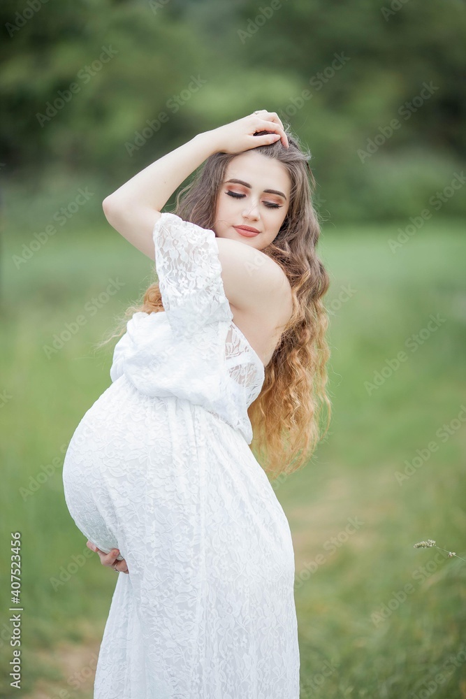 A beautiful young pregnant woman with curly long hair in a white dress walks in the field. Spring portrait of a pregnant woman. Happy pregnancy. A beautiful young pregnant woman with curly long hair in a white dress walks in the field. Spring portrait of a pregnant woman. Happy pregnancy.