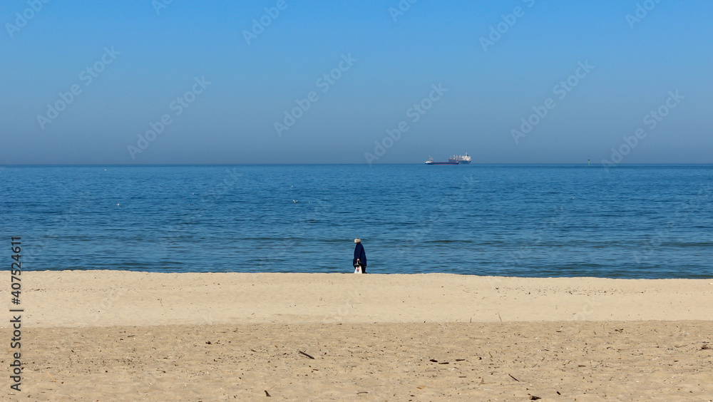 Filmic scene from seaside. Beautiful view of an old woman walking in the beach on a cold day and distant ships in the sea with a blue sky.