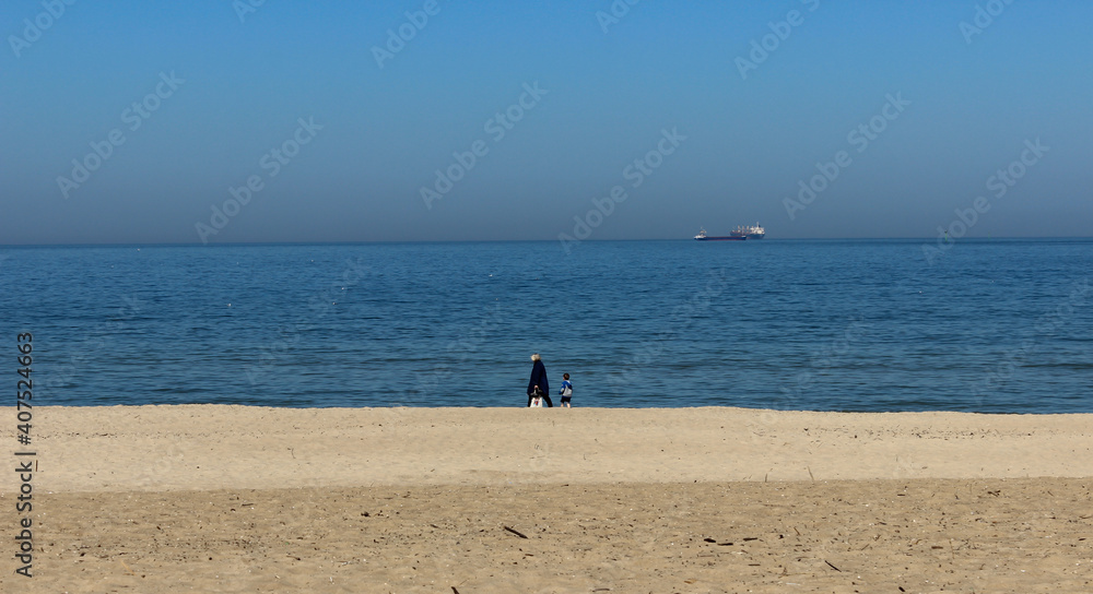 Filmic scene from seaside. Beautiful view of an old woman walking in the beach with her grandson on a cold day and distant ships in the sea with a blue sky.