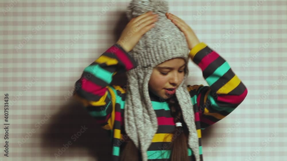 A beautiful dark-skinned little girl in a bright striped sweatshirt and a big winter hat with a bubo makes a grimace. Emotional happy child.