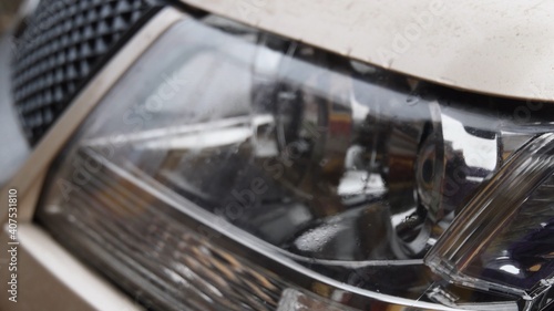 Car headlight wet after rain with blurry reflections in mirrored surface. Raindrops on dirty metallic car bonnet and blurred background